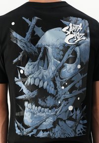 Zwarte T-shirt met een blauwe schedelafbeelding omringd door scherpe, textuurlijke vormen en witte accenten. Het ontwerp benadrukt diepte en contrast.