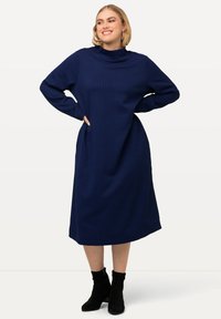 Marineblaues Langarmkleid mit hohem Kragen, das ein strukturiertes Muster aufweist, knielang ist und über Seitentaschen verfügt. Kombiniert mit schwarzen Stiefeletten.