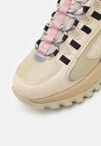 Beiger Sneaker mit strukturierter Sohle, rosa Schnürung, Mesh-Akzenten und grauen Details. Verfügt über eine abgerundete Spitze und mehrlagige Designelemente.