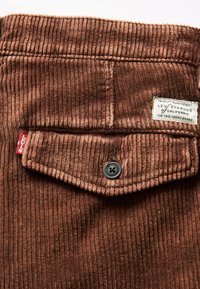 Pruunist korduroo kangast, millel on vertikaalsed sooned. Omab klapp-taskut, millel on must nupp ja valge brändi silt, millel on tekst "Levi Strauss". Osa on punasest sildist.