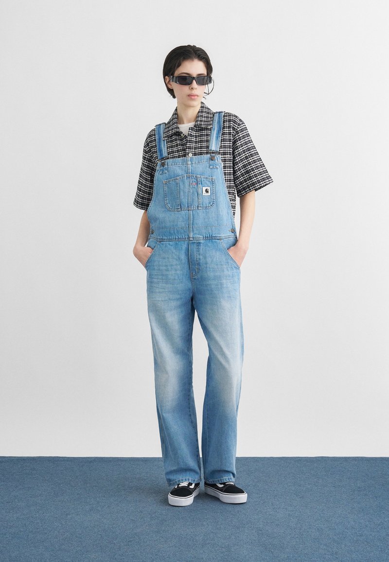 Personne portant une chemise à carreaux noir et blanc, une salopette en jean bleu clair, des lunettes de soleil noires et des baskets noires, debout sur un tapis bleu.