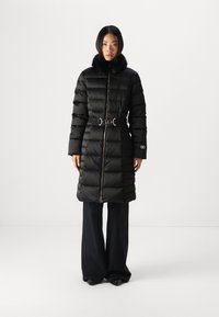 Zwarte pufferjas met een bontkraag, ceintuur in de taille, gequilt textuur en een rits aan de voorkant. Gecombineerd met ruime zwarte broeken en zwarte schoenen.