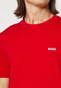 Personne portant un t-shirt rouge vif à manches courtes avec le mot "BOSS" imprimé en blanc sur la poitrine gauche.