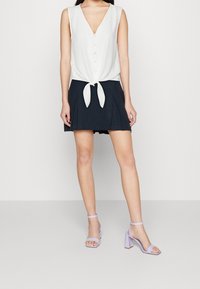 Blusa bianca senza maniche con scollo a V e legatura frontale; abbinata a una mini gonna a pieghe nera. Sandali con tacco a blocco lavanda completano l'outfit.