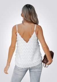 Witte mouwloze top met een diepe V-rug, met een textuurfranje ontwerp en dunne bandjes. Model houdt een bruine tas vast. Lichte denim jeans.