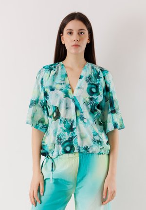 Giovane donna con lunghi capelli scuri indossa una camicetta floreale turchese e verde e pantaloni sfumati abbinati, in piedi davanti a uno sfondo semplice.