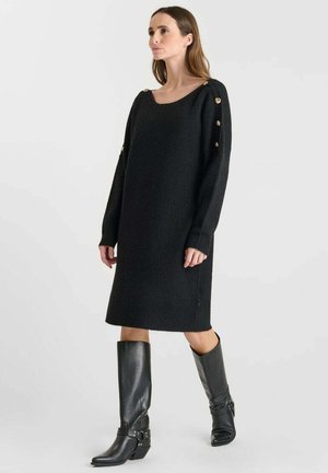 Robe pull noire en tricot avec une coupe ample, un large col et des boutons sur le côté. Associée à des bottes montantes noires à talons.