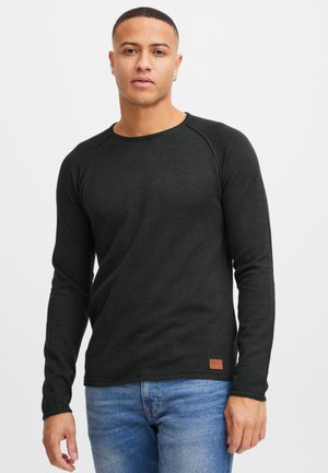 Mørk grå langermet t-skjorte i en ribbet tekstur, med rund utringning og en liten brun logo lapp nær kanten. Kombinert med blå jeans.