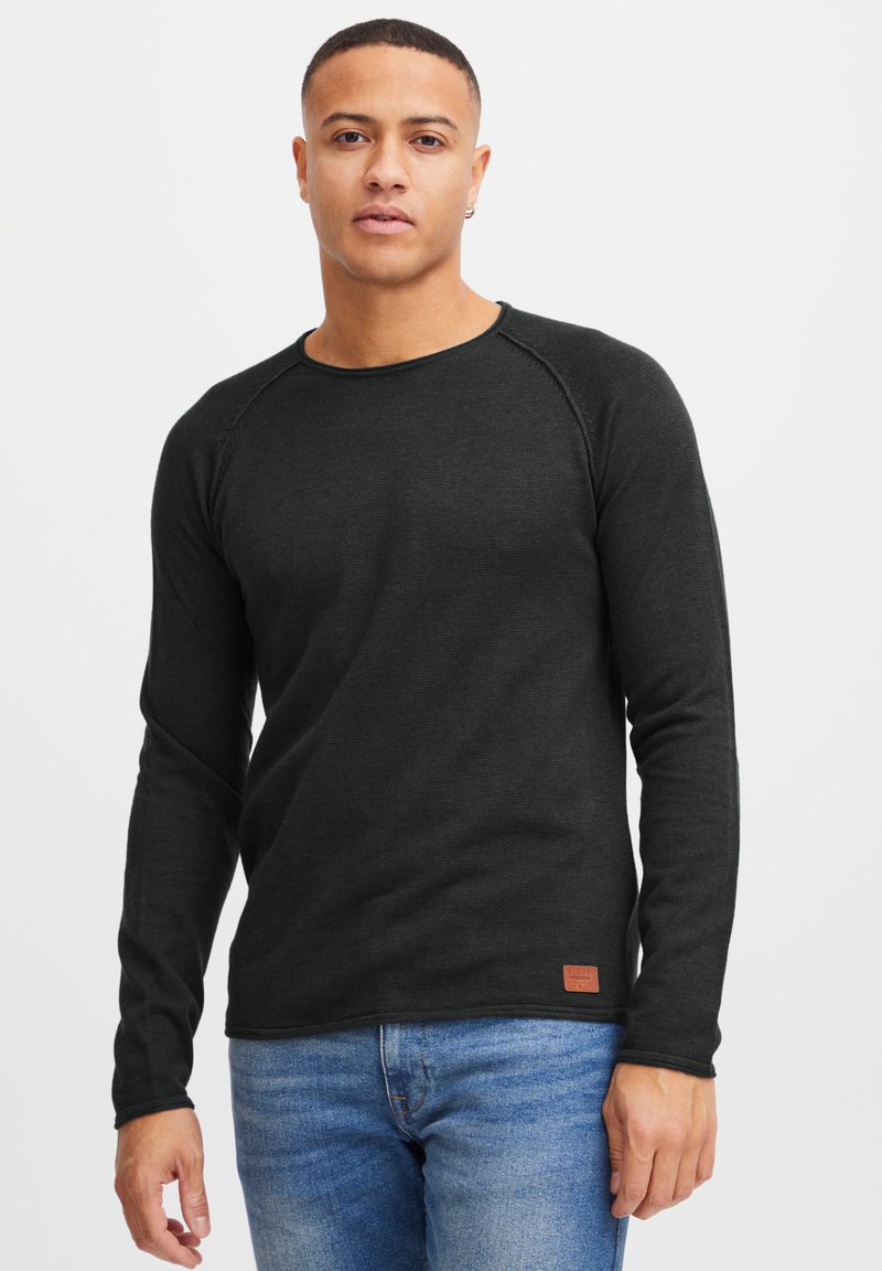 Mørk grå langermet t-skjorte i en ribbet tekstur, med rund utringning og en liten brun logo lapp nær kanten. Kombinert med blå jeans.