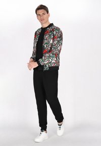 Giacca bomber con stampa floreale e fenicottero vivace, zip nera, abbinata a pantaloni jogger neri e scarpe da ginnastica bianche. Sfondo bianco.