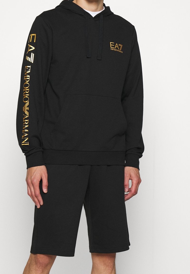 Hombre vestido con sudadera negra con capucha y pantalones cortos a juego, con el logo dorado "EA7 Emporio Armani" en el pecho y la manga, de pie contra un fondo blanco.