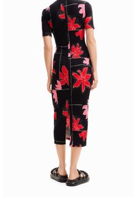 Robe en tricot noire ornée de grands motifs floraux rouges et roses, avec des accents de couture blanche et une fente sur le côté. Associée à des chaussures noires texturées.