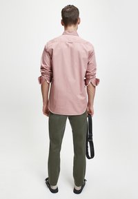 Camicia a maniche lunghe rosa chiaro con maniche arrotolate, abbinata a pantaloni slim fit verde oliva e sandali neri. Design semplice e casual.
