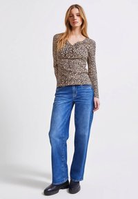 Vrouw staand in een beige langemouwen top met luipaardprint, blauwe jeans met rechte pijpen en zwarte enkellaarsjes, tegen een witte achtergrond.