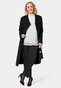 Schwarzer Wollmischungs-Mantel mit offenem Vorderteil, texturiertem Design, getragen über einer weißen Bluse und schwarzen Hosen, kombiniert mit spitzen schwarzen Absatzschuhen.