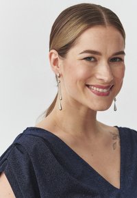 Strieborné visacie náušnice visia z obručového kruhu a majú elegantný dizajn s predĺženými slzovitými tvarmi. Modelka má na sebe tmavomodrý top.