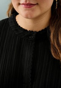 Schwarze Bluse mit Struktur und Wellenkragen, ausgestattet mit einem einzigen schwarzen Knopfverschluss an der Vorderseite und gefältelten Stoffdetails.