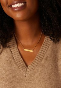 Gouden balkhalsketting aan een fijne ketting, met een rechthoekige hanger tegen een gestructureerde, tan kleurige trui met een V-hals.