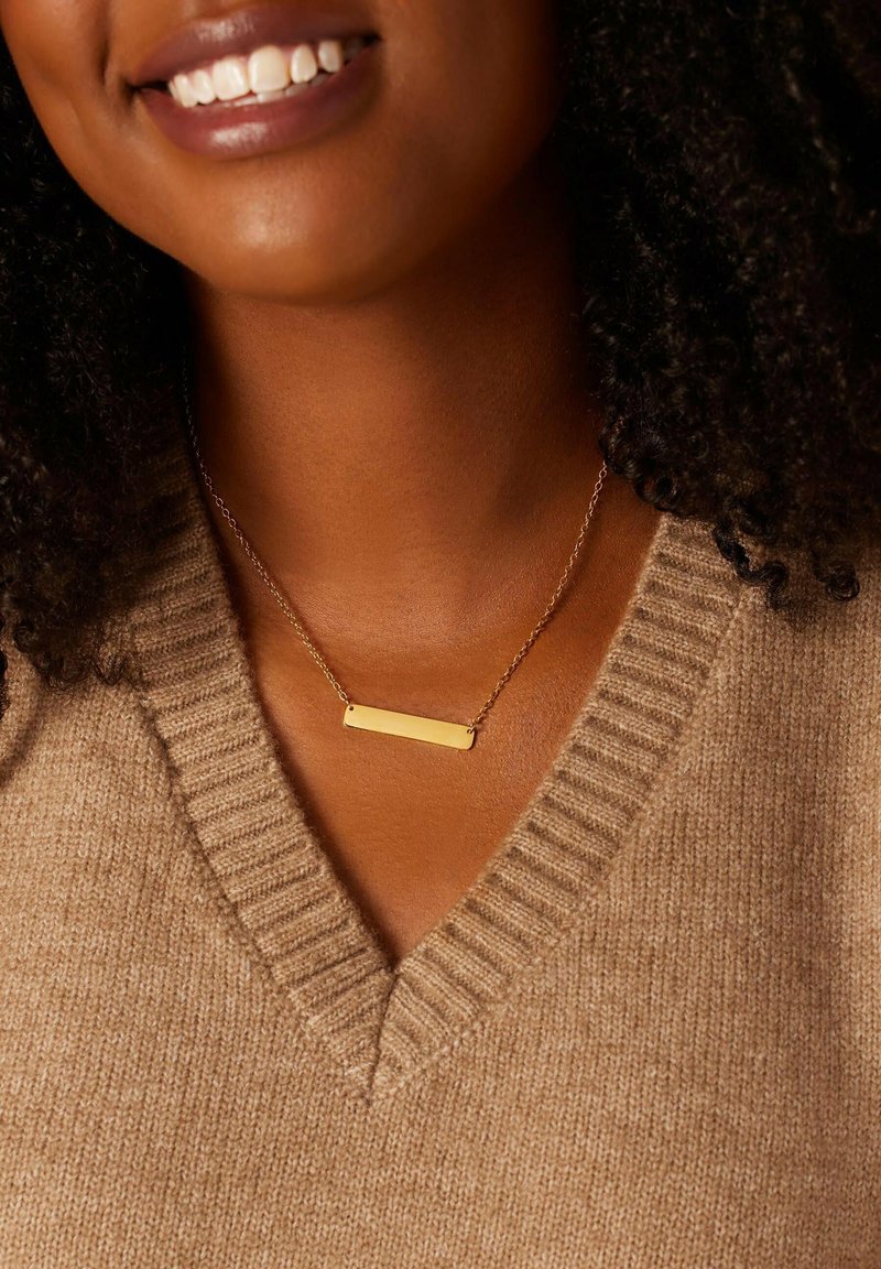 Gouden balkhalsketting aan een fijne ketting, met een rechthoekige hanger tegen een gestructureerde, tan kleurige trui met een V-hals.