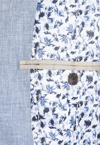 Modrý a bílý textil s květinovým vzorem a strukturovanými herringbone sekcemi, doplněný béžovou stuhou a tmavým knoflíkovým detailem.