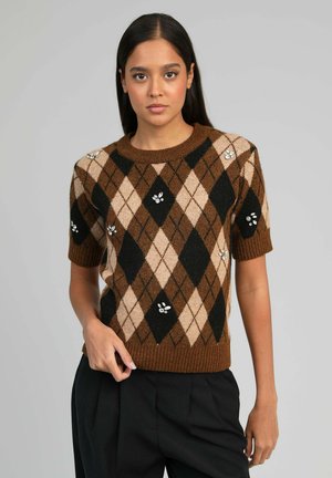 Jeune femme portant un pull en tricot à manches courtes à motif losanges marron et beige avec des ornements de bijoux, assorti à un pantalon noir.