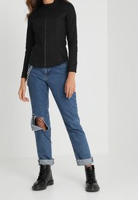 Svart zip-up jacka med hög krage, som kombineras med ljusblå distressed jeans med ett stort hål och uppvikta muddar. Svarta snörskor.