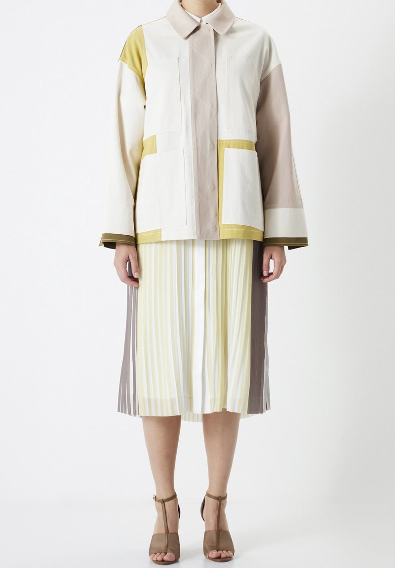 Veste multicolore en beige, vert et marron, avec de grandes poches et un col. Associée à une jupe plissée rayée jaune et blanche.