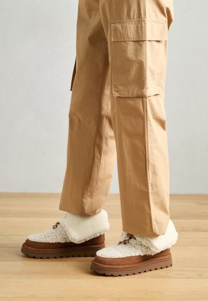 Weiße und braune, flauschige Schuhe mit dicker Sohle, die einen weichen Bund und Schnürdetails aufweisen, kombiniert mit hellbraunen Cargo-Hosen.