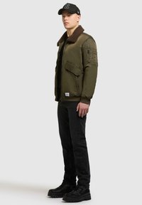 Olivgrüne Bomberjacke mit braunem Fellkragen, Fronttaschen und geschichteter Gestaltung, kombiniert mit schwarzen Jeans und schwarzen Stiefeln.