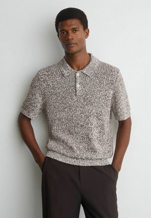 Mann trägt ein kurzärmeliges, strukturiertes Strick-Poloshirt mit zwei Knöpfen und dunkle Hose, steht mit den Händen in den Taschen vor einer schlichten Wand.