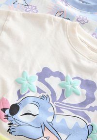 Dwie koszulki ułożone jedna na drugiej. Jedna w kolorze kremowym z teksturowanymi kwiatami z filcu i niebieską postacią z kreskówki; druga w jasnoniebieskim z kwiatowymi wzorami.