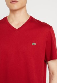T-shirt rossa in cotone con scollo a V, caratterizzata da un piccolo logo di un coccodrillo ricamato di colore verde sul lato sinistro del petto. Tessuto morbido.