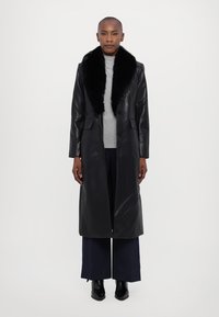 Μαύρο δερμάτινο μακρύ παλτό με ψεύτικη γούνα στη воротά, δύο μπροστινές τσέπες και ίσια γραμμή σχεδίασης, φορεμένο πάνω από γκρι μπλούζα και παντελόνι με φαρδιά πόδια.