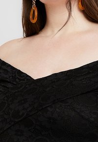 Svart spetsklänning med off-shoulder-design och blommönster. Tyget har en texturerad yta och lagerdetaljer.