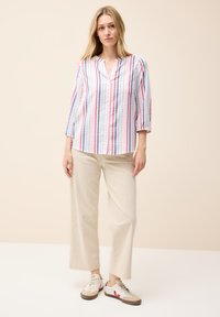 Camicia a righe bianca, rossa e blu, con scollatura a V e maniche a tre quarti. Abbinata a pantaloni larghi beige e sneaker.