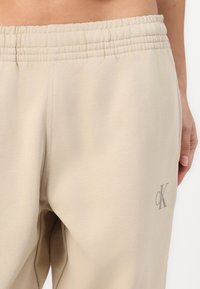 Calças de fato beje com um cós elástico, textura macia e um pequeno logótipo bordado em cor contrastante perto da costura lateral.