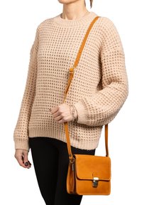 Borsa a tracolla in pelle color tan con chiusura a pattina e finiture in metallo, abbinata a un maglione di lana beige e pantaloni neri.