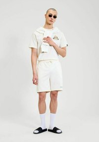 Hombre vestido con un atuendo casual color blanco roto, que incluye pantalones cortos, una camiseta, un suéter colocado sobre los hombros, calcetas blancas, chanclas negras y gafas de sol.