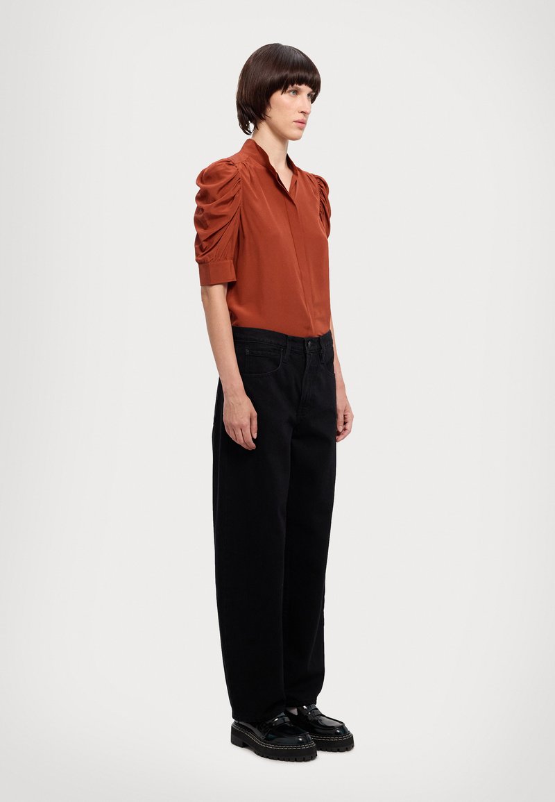 Blusa de cor rustica com mangas volumosas, com abertura de botões; combinada com calças pretas de corte solto e sapatos de plataforma pretos.