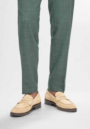 Bežīgas zamšādas loferes ar zemu papēdi, smalkām šuvēm un platām siksnām, saderīgas ar pieguļošām zaļām teksturētām biksēm.