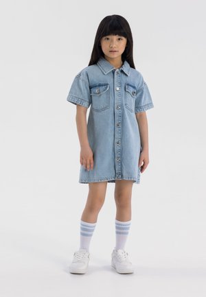 Robe en denim à manches courtes, fermeture par boutons sur le devant, deux poches poitrine, couleur bleu clair et coupe légèrement décontractée. Portée avec des baskets blanches.