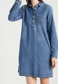 Denimová košile šaty s límečkem, dlouhými rukávy, jedním náprsním kapsou a bočními rozparky; střední modrá barva s jemnými švy.