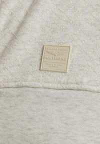 Tissu matelassé gris clair présentant un motif de diamond texturé. Une étiquette beige avec un texte en relief et des ailes est positionnée en bas à droite.