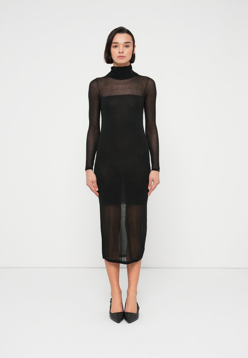 Γυναίκα που στεκεται μπροστά σε λευκό φόντο, φορώντας μια διάφανη μαύρη midi φόρεμα με μακριά μανίκια και ζιβάγκο, και μαύρες μυτερές γόβες.