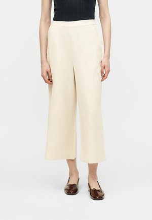 Personne portant un pantalon beige taille haute à jambes larges, court au-dessus des chevilles, avec des ballerines brillantes marron foncé, sur un fond uni.