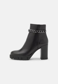 Bottine en cuir noir à talon épais, semelle texturée et accent en chaîne argentée autour de la cheville. Design épuré avec un minimum de détails.