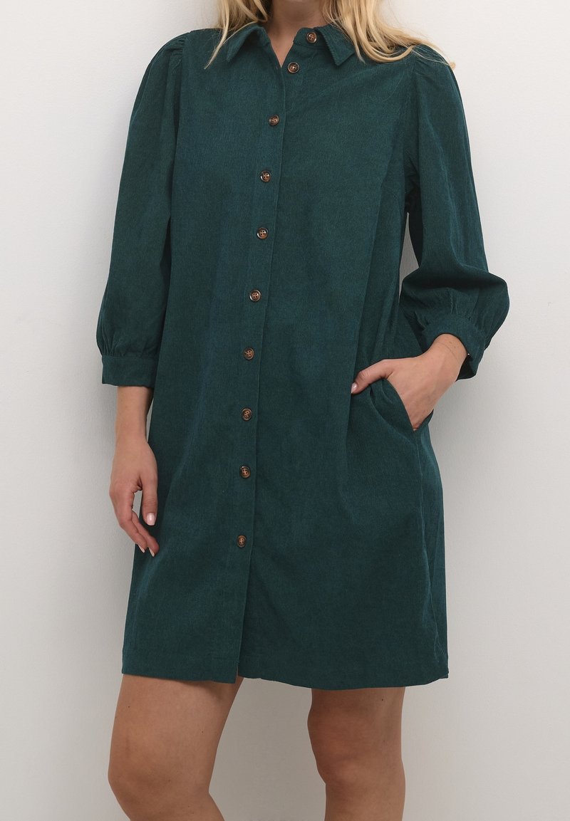 Femme portant une robe boutonnée vert foncé avec des manches ballon trois-quarts et une main dans la poche latérale.