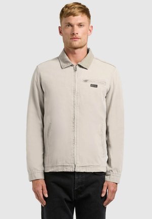 Muž so svetlohnedými krátkymi vlasmi, oblečený v svetlo béžovej zipsovej bunde s golierom a čiernych nohaviciach, čelom k prednej strane proti jednoliatemu pozadiu.