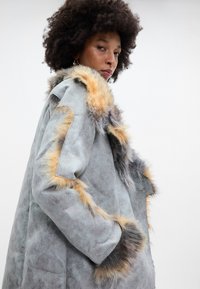 Pelēkas zamšādas mētelis ar daudzkrāsainu mākslīgā kažokāda apdari gar apkakli un pleciem, ar brīvu piegriezumu un sānu kabatām.