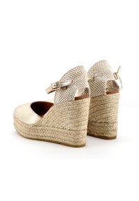 Espadrillas a zeppa color oro con cinturino intrecciato nella parte posteriore, suola in corda di juta e materiale superiore liscio. Presenta una fibbia regolabile alla caviglia.